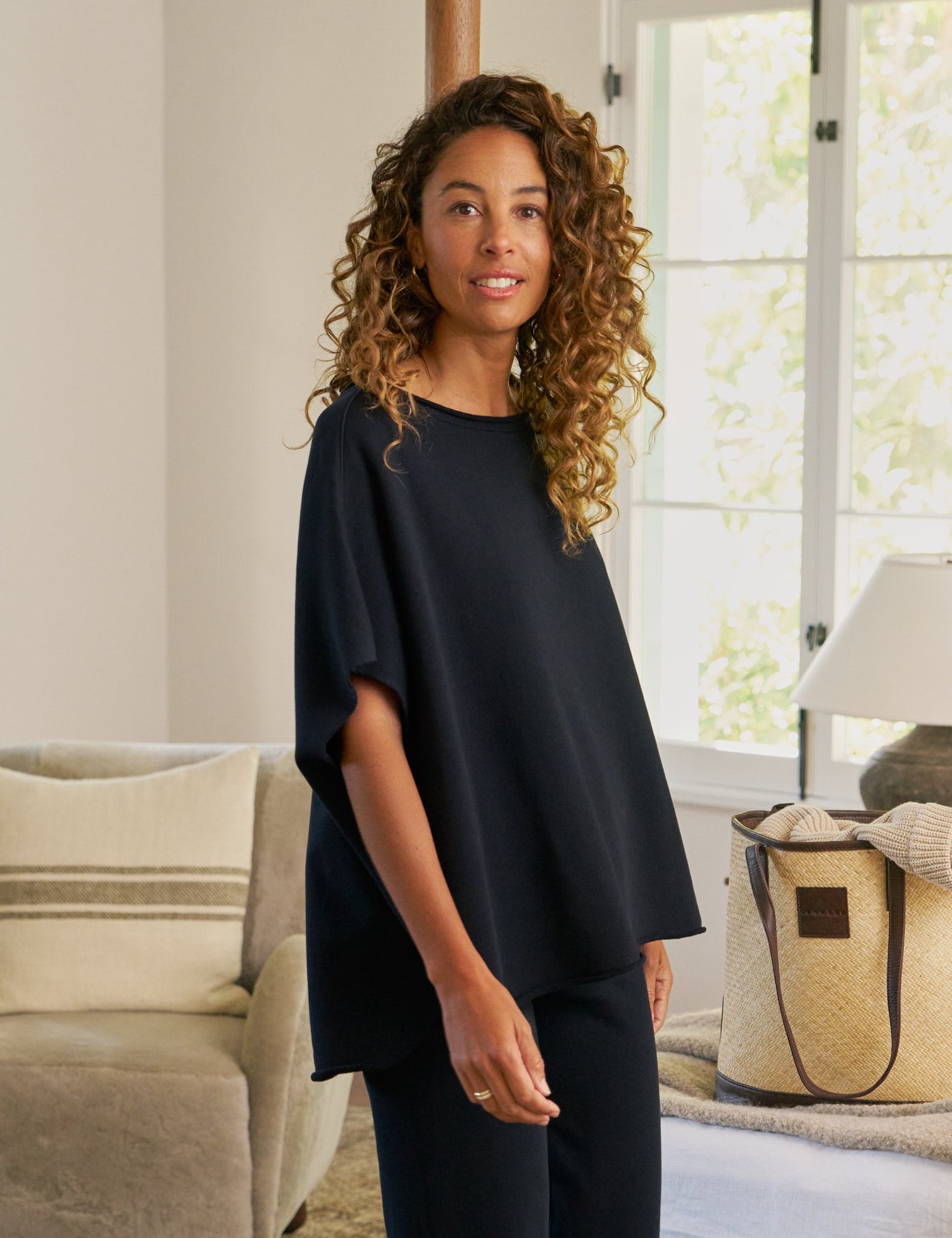 Woman in a navy outfit standing in a bedroom with a bed and lamp in the background., alternate view