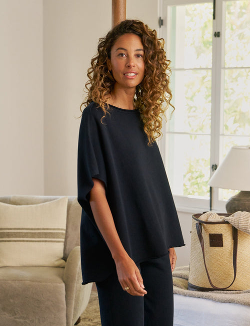 Woman in a navy outfit standing in a bedroom with a bed and lamp in the background., alternate view