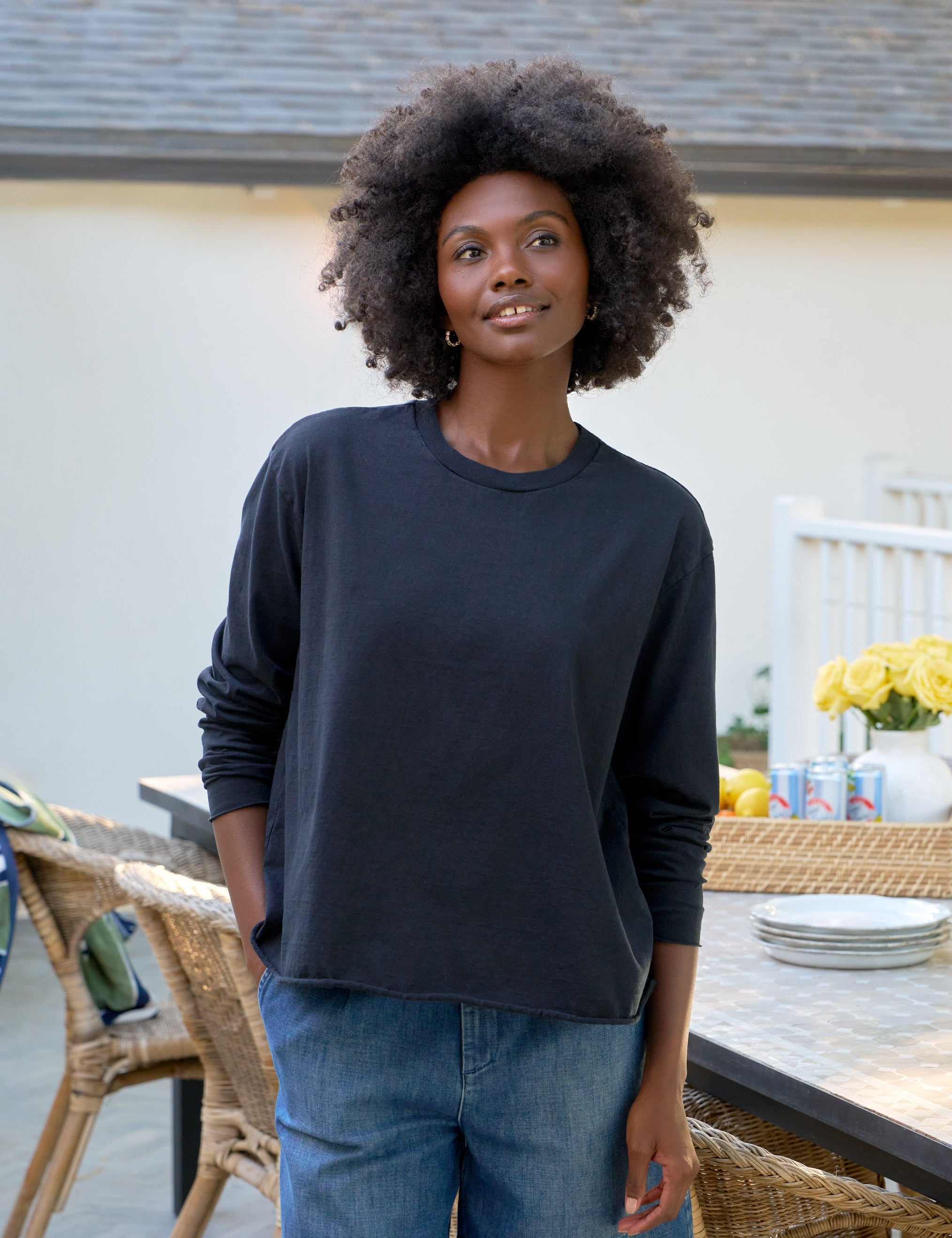 Woman standing outdoors in a casual setting with furniture and decor.