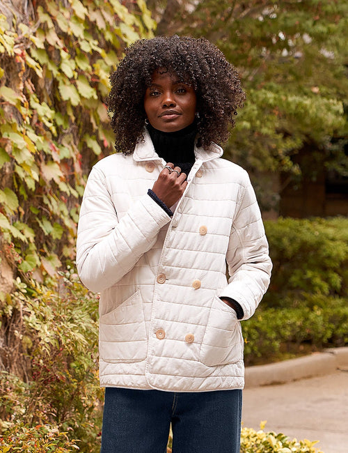 A woman stands outdoors on a stone path wearing a cream quilted double-breasted jacket, navy wide-leg trousers, and black shoes, with ivy-covered trees and shrubs in the background., front view
