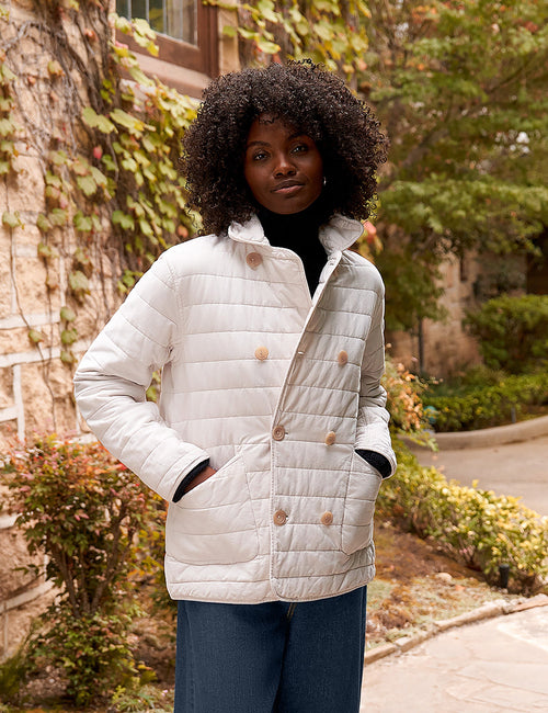A woman wearing a light cream quilted jacket with brown buttons, a black turtleneck, and dark blue wide-leg jeans, standing outdoors in front of a stone wall with ivy., view 7