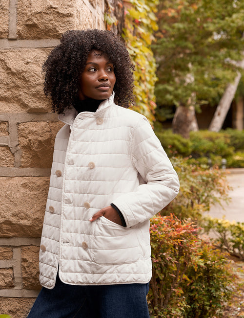 A woman with dark curly hair wearing a cream quilted jacket and dark blue pants, standing outdoors next to a stone wall with green foliage in the background., view 3