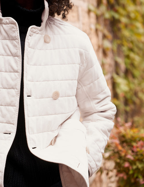 A close-up of a person wearing a light cream quilted jacket over a black sweater, with a blurred outdoor background of foliage and a stone wall., view 5