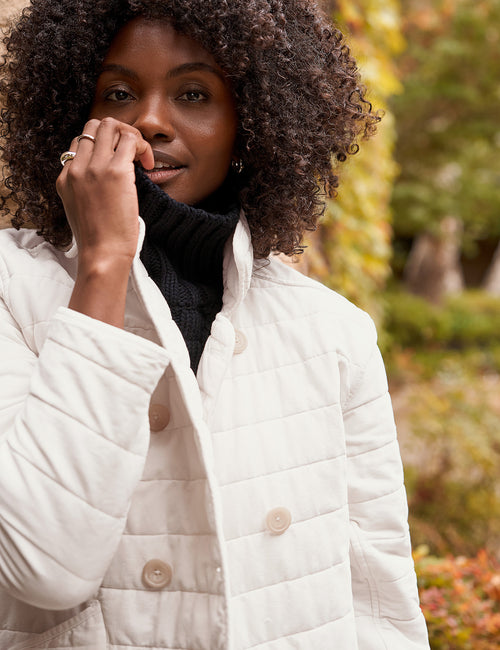 A woman with curly dark hair wearing a cream quilted jacket over a black turtleneck, standing outdoors against a blurred background of autumn foliage., view 6