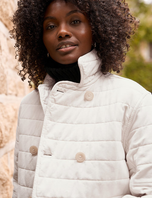 A close-up shot of a woman wearing a cream-colored quilted jacket with a high stand-up collar and beige buttons, set against a soft-focus outdoor background., alternate view