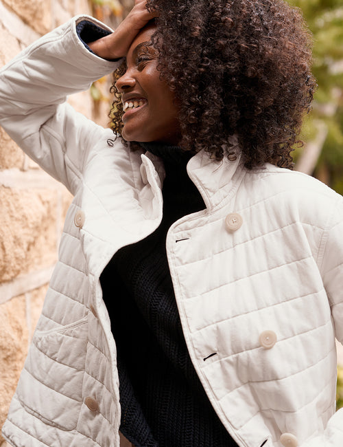 A woman wearing a cream quilted jacket over a black top, standing outdoors against a stone wall., view 4
