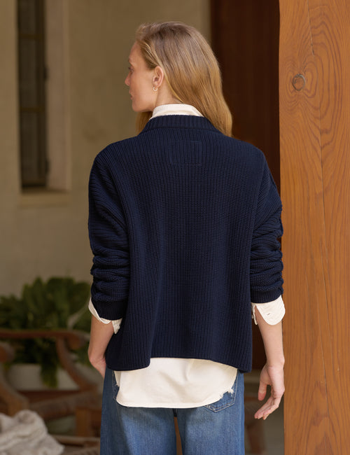 A woman shown from the back wearing a dark navy ribbed knit cardigan over a cream shirt and blue jeans, standing indoors near a wooden door., alternate view