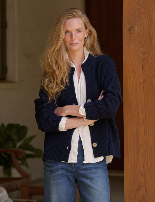 A woman with long blonde hair stands indoors wearing a navy ribbed cardigan over a white button-up shirt and blue jeans, with her arms crossed., front view