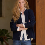 A woman with long blonde hair stands indoors wearing a navy ribbed cardigan over a white button-up shirt and blue jeans, with her arms crossed.