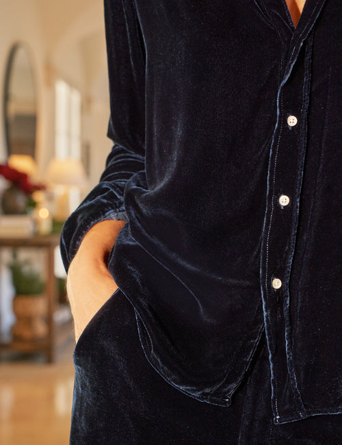 A close-up of a person wearing a dark navy velvet button-front shirt with a relaxed fit, standing in a softly blurred indoor room., alternate view