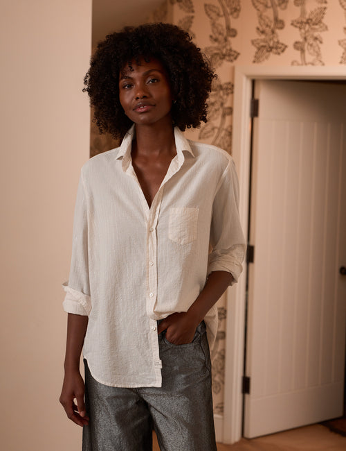 A woman with curly black hair wearing a white long-sleeve button-down shirt with rolled-up sleeves and gray pants, standing in a doorway with patterned wallpaper in the background., front view