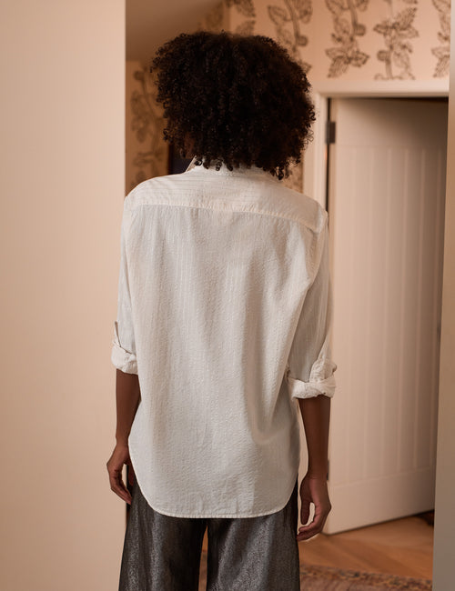 A person viewed from behind wearing a loose white button-up shirt with rolled sleeves and dark gray pants in an indoor room with patterned wallpaper and a white door., alternate view