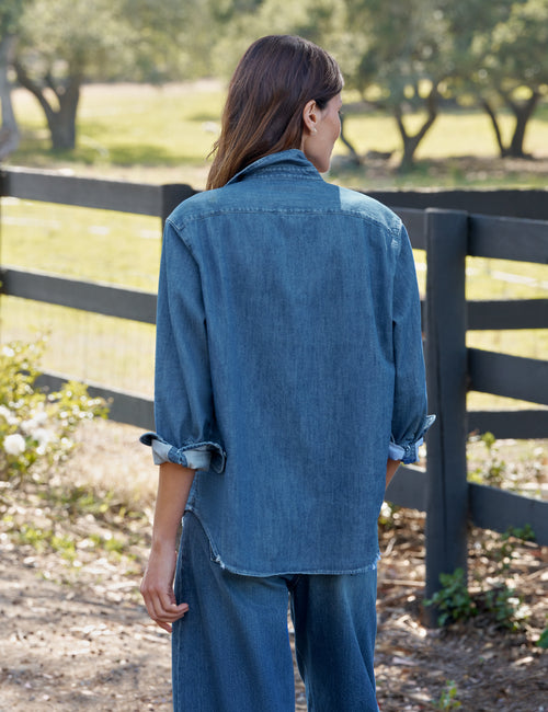 A woman viewed from behind wearing a blue denim shirt and matching wide-leg jeans, standing outdoors in front of a dark wooden fence with trees in the background., alternate view