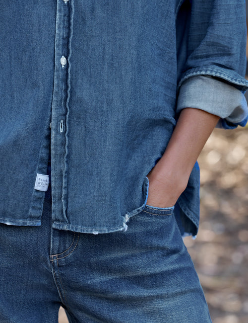 Close-up of a person wearing a blue denim button-up shirt with rolled sleeves and matching blue denim jeans, with one hand in a pocket., view 3