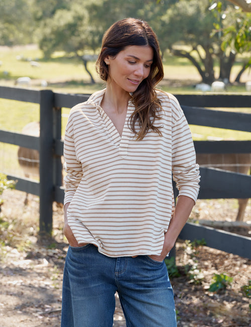 A woman wearing a beige and brown striped long-sleeve collared shirt with blue jeans, standing outdoors in front of a wooden fence with a blurred farm background., view 4