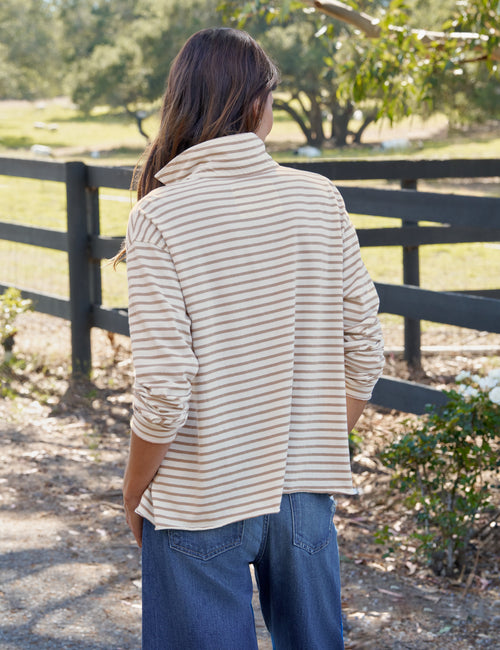 A woman viewed from behind wearing a cream and tan horizontally striped long-sleeve shirt and blue jeans, standing outdoors in front of a black wooden fence and trees., alternate view