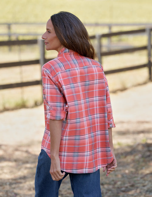 A woman wearing a coral red and blue plaid button-down shirt with rolled-up sleeves, paired with dark blue jeans, standing outdoors in front of a wooden fence., alternate view