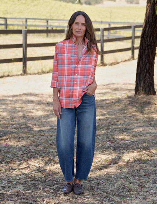 A woman standing outdoors in a rural setting, wearing a coral red and blue plaid button-down shirt, wide-leg blue jeans, and dark brown loafers., view 5