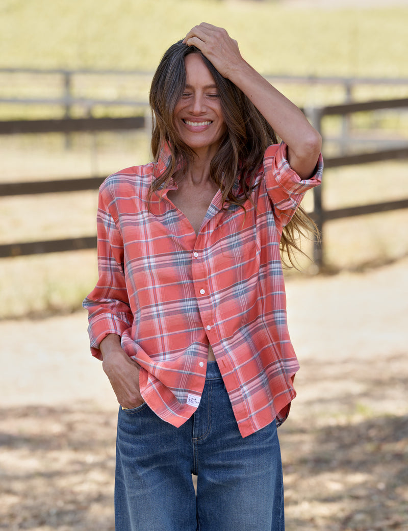 A woman wearing a coral red and blue plaid button-down shirt with rolled-up sleeves, paired with blue jeans, standing outdoors in front of a wooden fence.