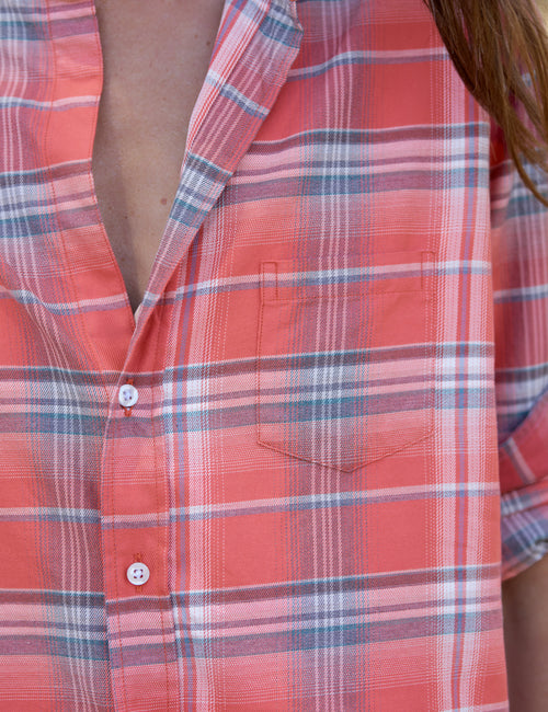 Close-up of a plaid short-sleeve button-up shirt worn by a model, featuring a coral red base with blue and white stripes., view 3