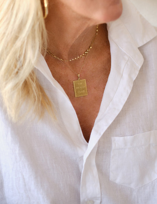 A close-up of a person wearing layered gold necklaces, including a square pendant engraved with 'THE BEACH CLUB' and a delicate beaded chain, paired with a white collared shirt., view 3