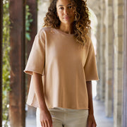 A woman with curly brown hair wearing a light brown-beige short-sleeve top and white pants, standing in a stone colonnade.