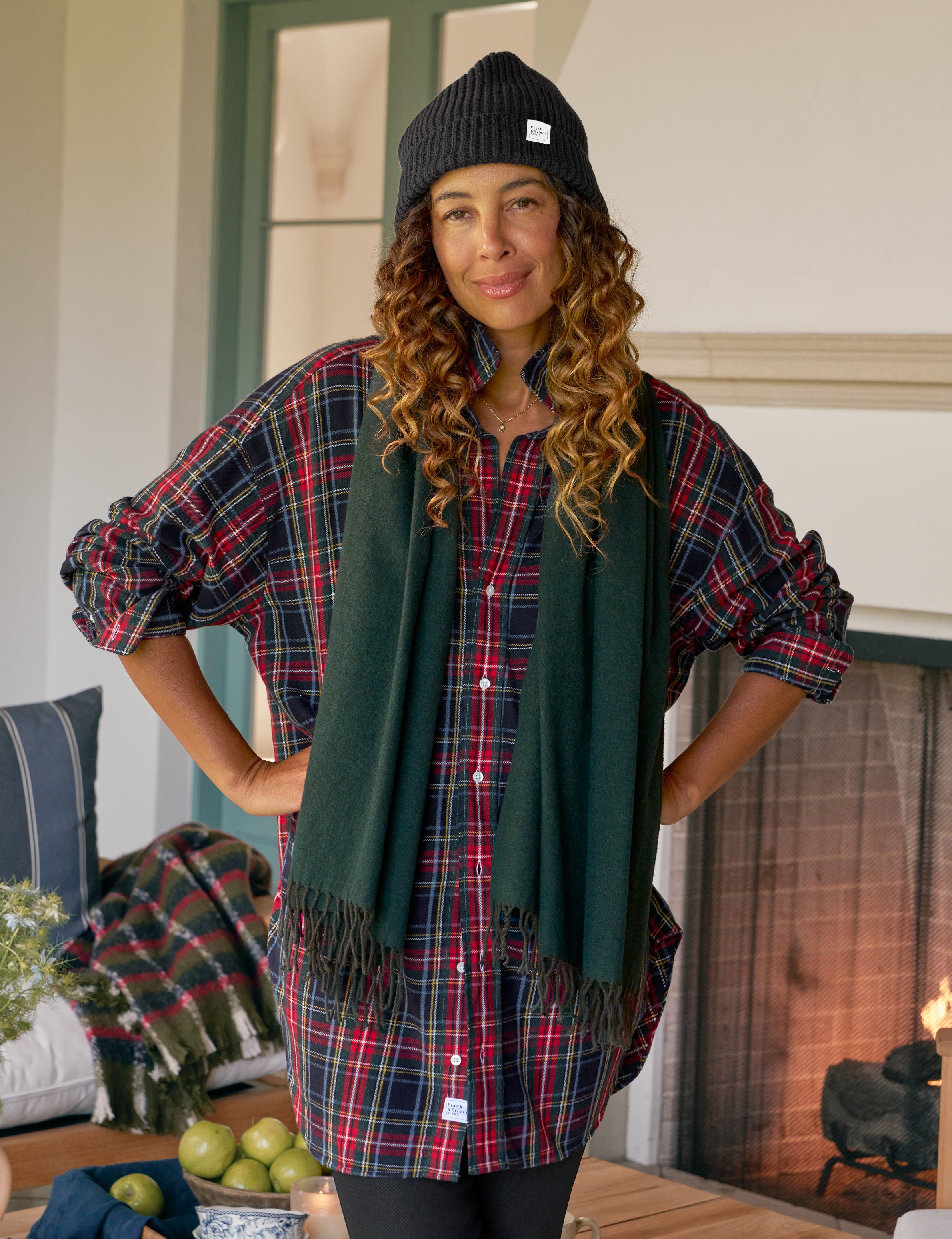 Woman wearing a plaid shirt, green scarf, and black beanie in a cozy indoor setting.