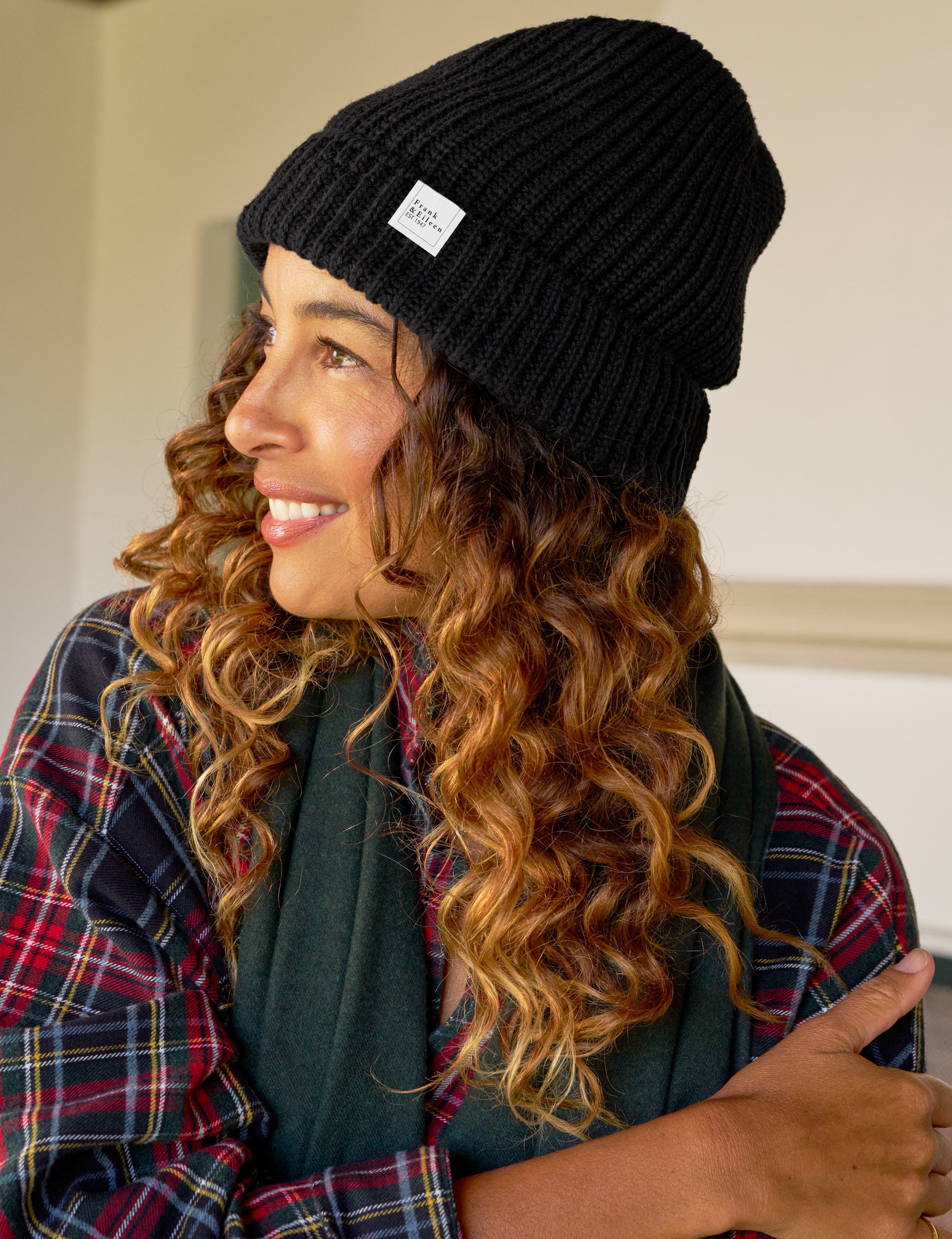 Woman wearing a black beanie with a logo, plaid shirt, and green scarf indoors.