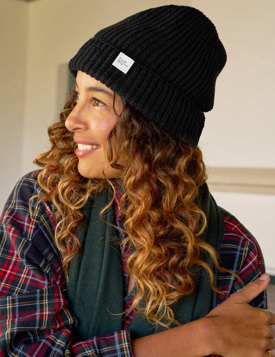 Woman wearing a black beanie with a logo, plaid shirt, and green scarf indoors.