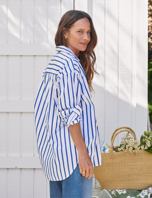 Woman wearing a blue and white striped shirt with rolled-up sleeves, standing next to a white fence., alternate view