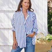 Woman wearing a blue and white striped shirt and jeans, standing next to a woven basket with flowers against a white wooden fence.