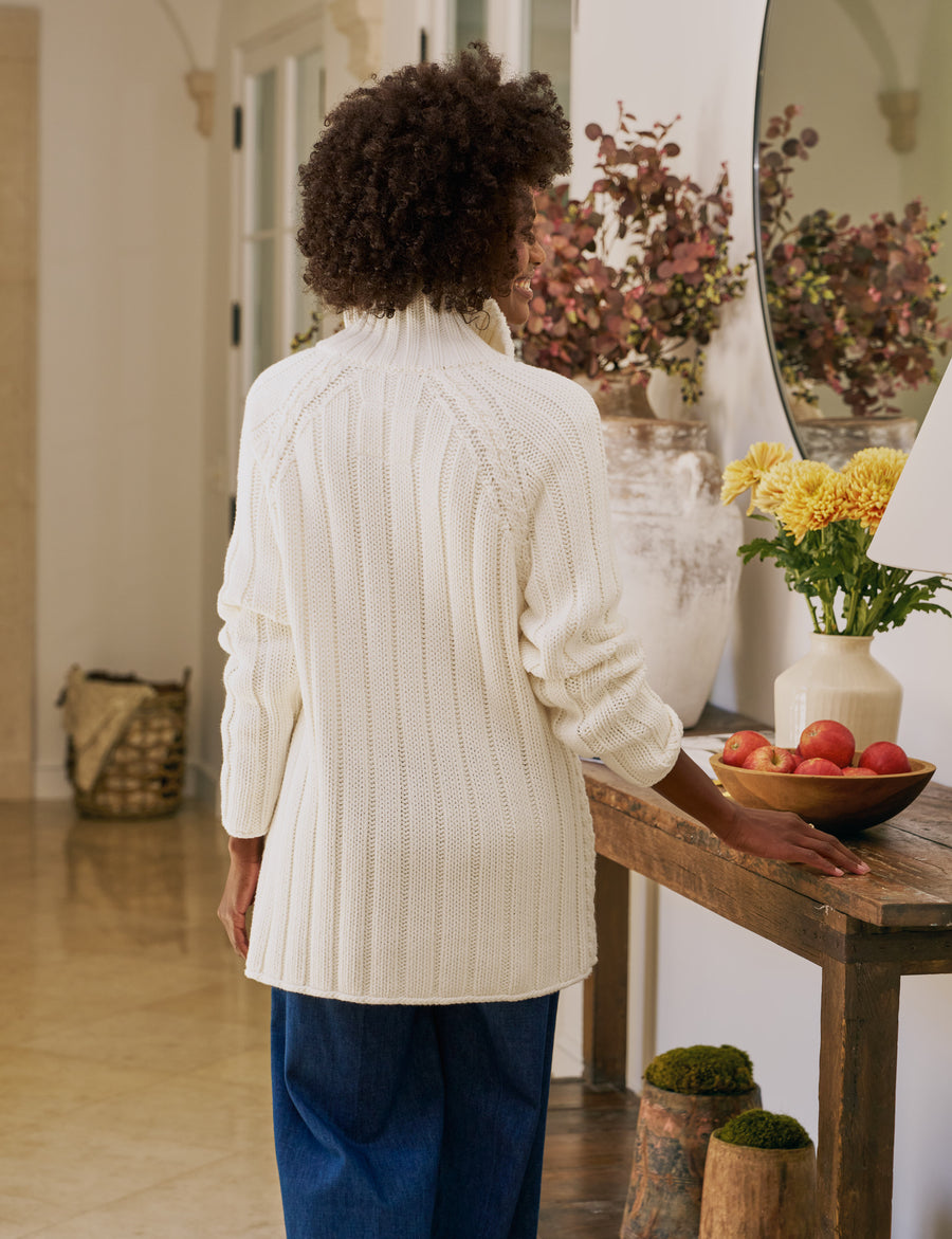 Person wearing a white ribbed sweater in a home setting with decor elements.