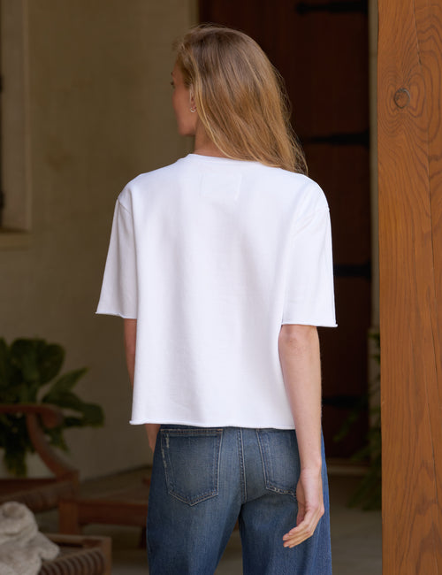 A woman viewed from behind wearing a white short-sleeve cropped T-shirt and blue jeans in an indoor setting with a wooden door frame., alternate view