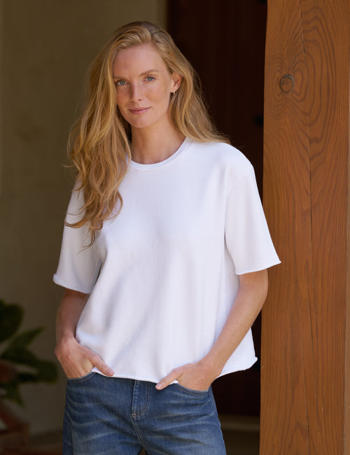 A woman with long blonde hair wearing a white short-sleeve crewneck t-shirt and blue jeans, standing in front of a wooden door., front view