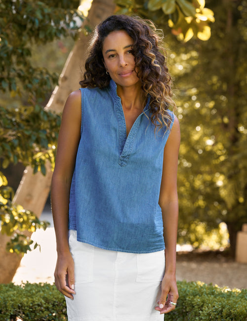 A woman wearing a sleeveless blue denim top with a split V-neck and white skirt, standing outdoors in front of a blurred background of green trees and foliage., front view