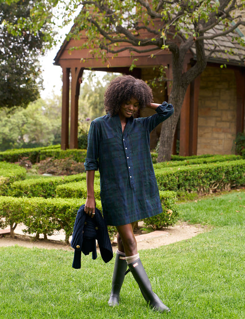 A woman stands on a grassy lawn in front of a wooden gazebo and trees, wearing a dark blue plaid shirt dress, tall black rain boots, and holding a dark jacket., view 4