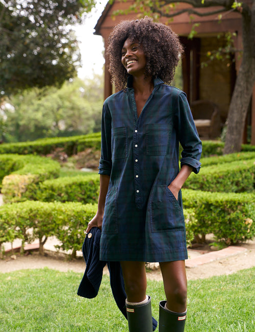 A woman outdoors in a garden wearing a dark navy plaid shirt dress with front pockets and buttons, holding a matching navy jacket, and wearing green rain boots., front view