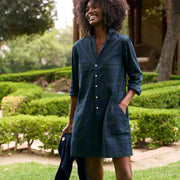 A woman outdoors in a garden wearing a dark navy plaid shirt dress with front pockets and buttons, holding a matching navy jacket, and wearing green rain boots.