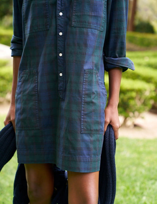 A person outdoors wearing a dark blue and green plaid shirt dress with a button front and large front patch pockets, holding a dark knit sweater in a grassy garden setting., view 3