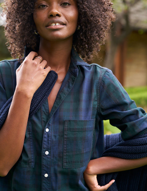 A close-up lifestyle shot of a woman wearing a dark teal and navy plaid button-down shirt with a knit cardigan draped off her shoulders., alternate view