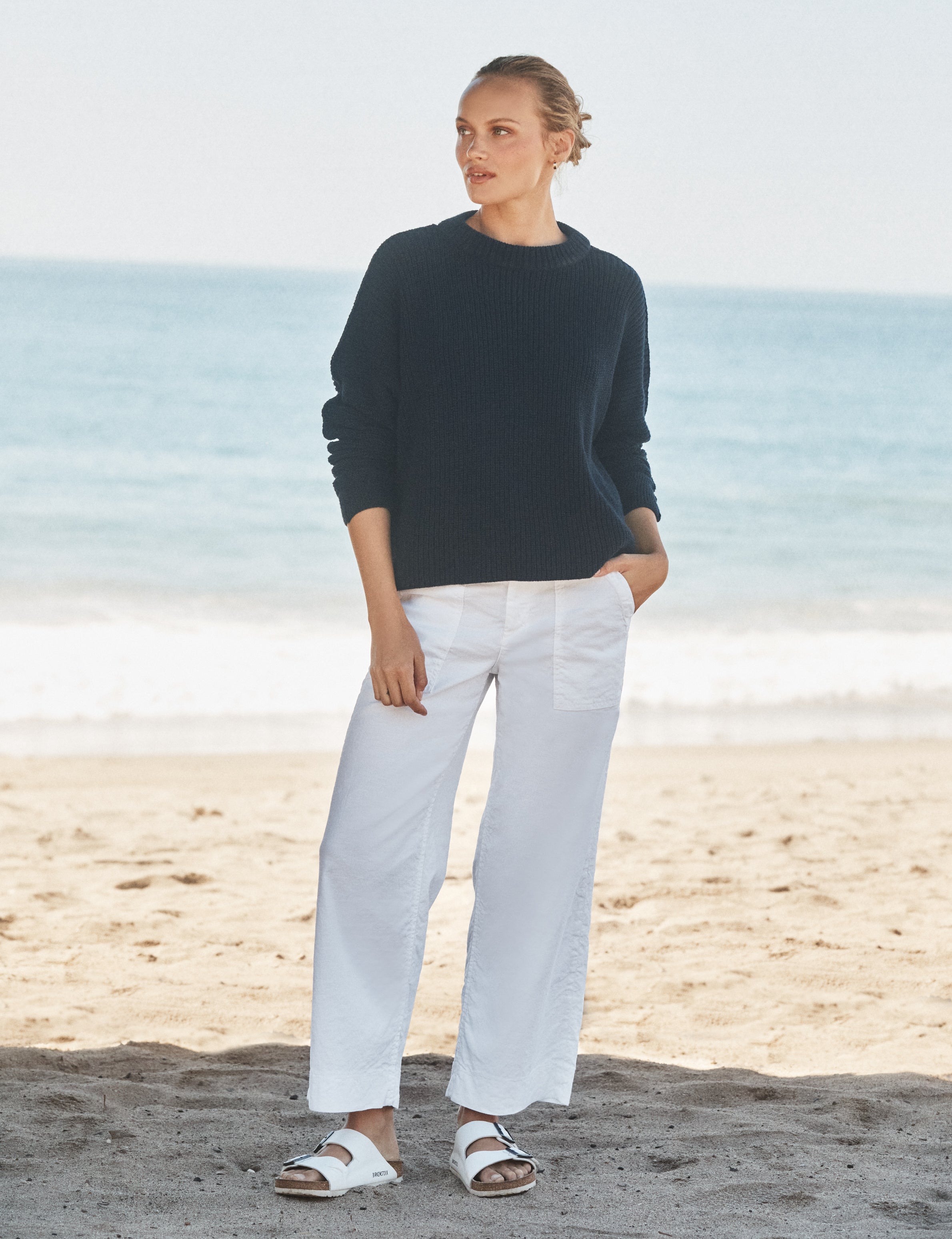 Woman standing on a beach wearing a navy sweater and white pants., view 4