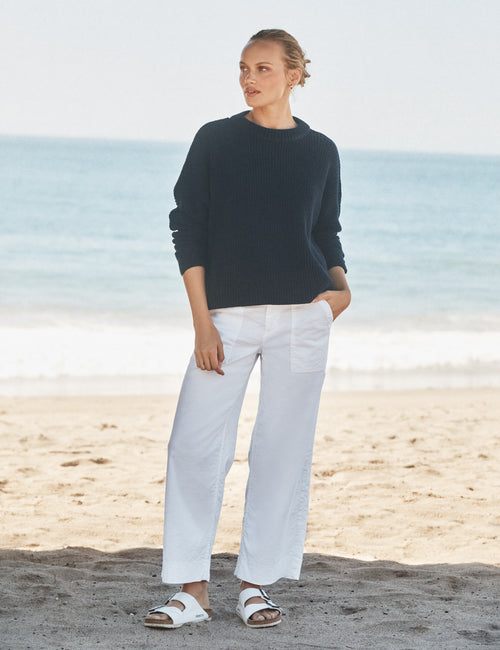 Woman standing on a beach wearing a navy sweater and white pants., view 4
