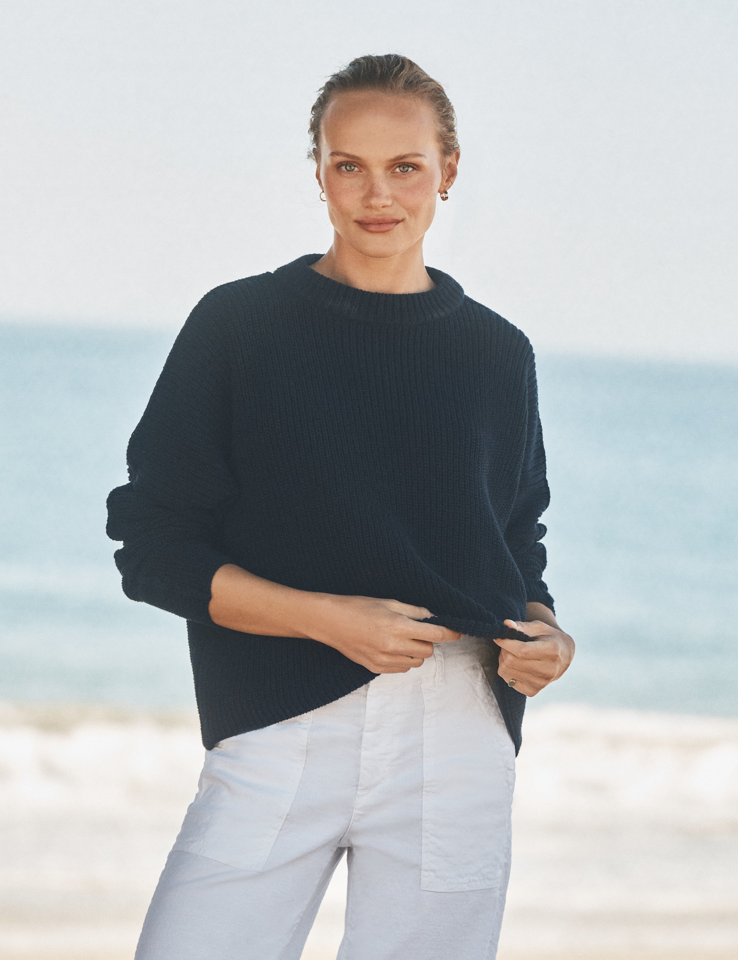 Woman wearing a navy sweater and white pants standing on a beach., front view