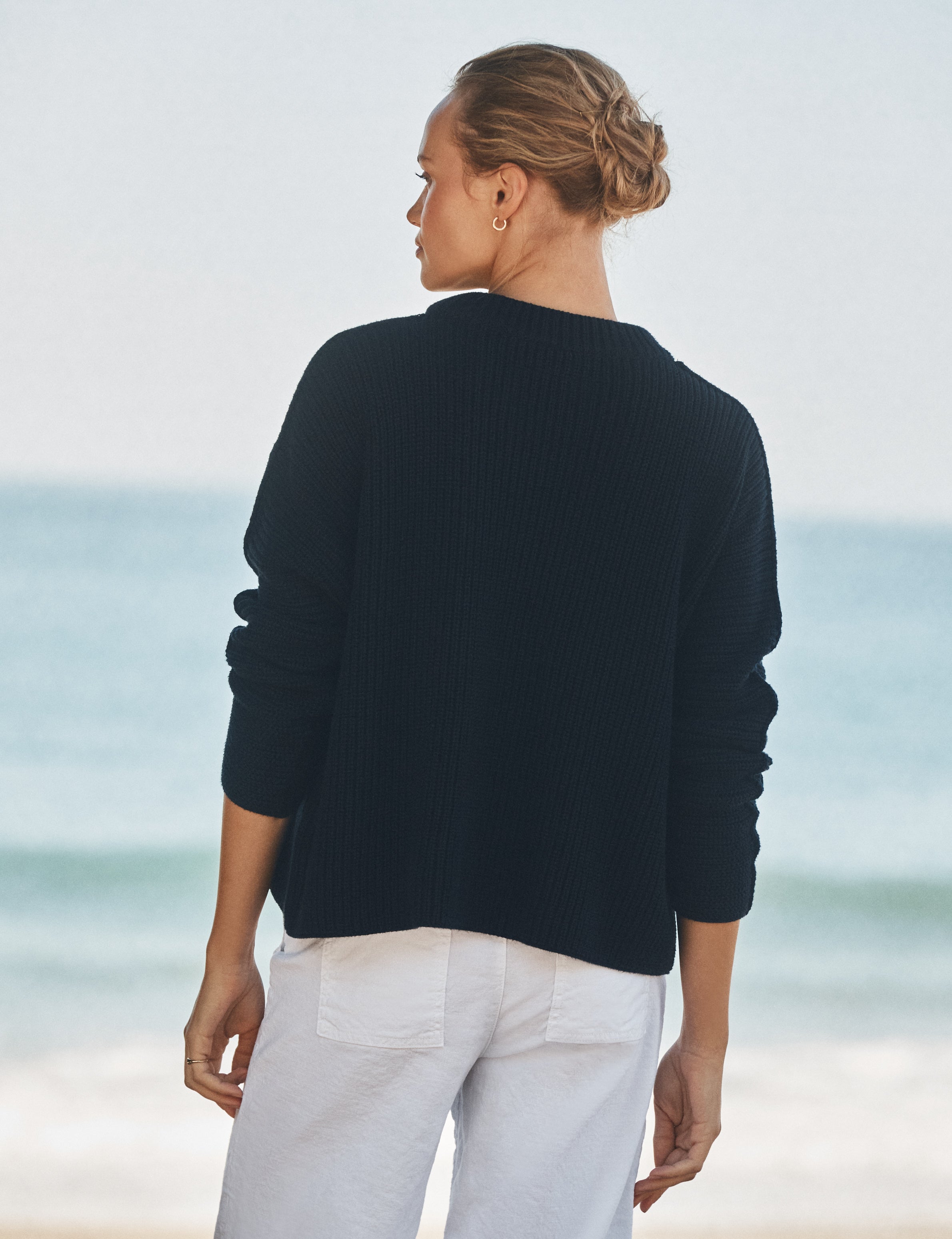 Person wearing a navy sweater and white pants standing on a beach with ocean view, alternate view