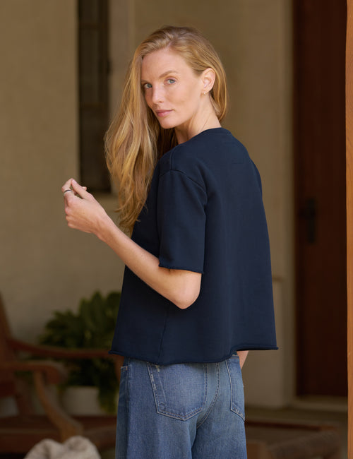 A woman with blonde hair is shown from the back wearing a dark navy blue short-sleeve top and blue jeans, standing in a warm-toned indoor setting., alternate view