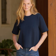 A woman with blonde hair wearing a dark navy blue short-sleeve top and blue jeans, standing in front of a wooden door.