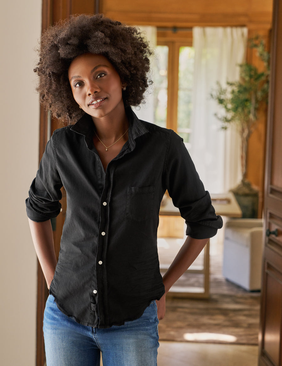 Woman wearing a black shirt and blue jeans standing in a room with wooden furniture and plants.