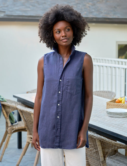 Woman wearing a sleeveless blue shirt standing outdoors near a table with chairs., front view