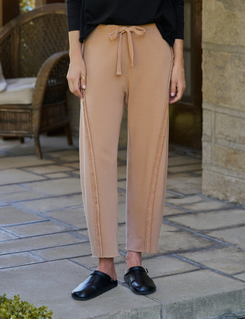 A person wearing tan drawstring pants with side seam detailing, paired with a black top and black loafers, standing on a stone patio., front view