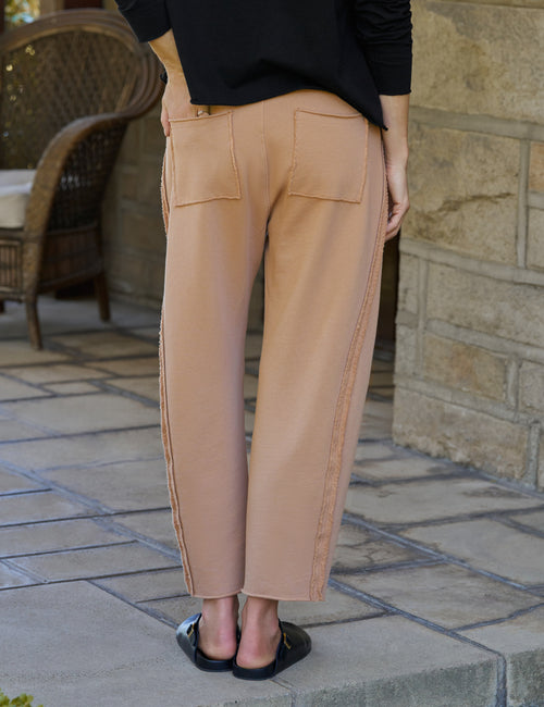 A person shown from the back wearing tan wide-leg cropped pants with side seam detailing, a black long-sleeve top, and black slide clogs, standing on a stone patio., alternate view